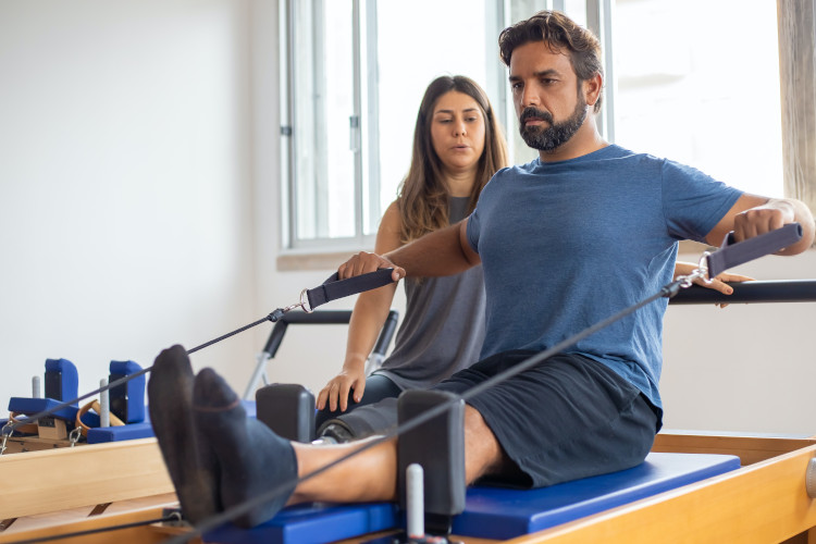 Ein Mann sitzt auf einem Pilates-Gerät und führt eine Übung mit Seilen aus, während eine Frau im Hintergrund zuschaut.